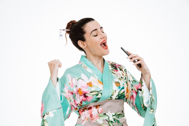 Glückliche und aufgeregte Frau im traditionellen japanischen Kimono, die Smartphone hält und es als Mikrofon verwendet, um ein Lied zu singen, das Spaß auf Weiß hat