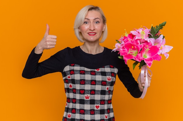 Kostenloses Foto glückliche und aufgeregte junge frau im schönen kleid, das blumenstrauß hält, der vorne lächelnd zeigt, zeigt daumen hoch, der internationalen frauentag steht über orange wand