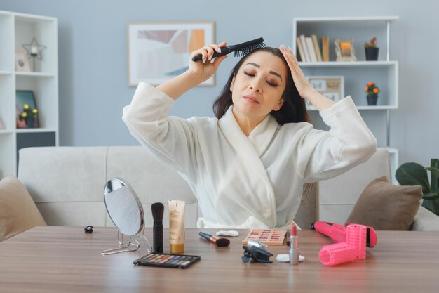 Glückliche und entspannte junge asiatin mit handtuch im bademantel, die zu hause am schminktisch sitzt und ihr haar berührt, das ihr haar bürstet und morgens make-up-routine macht