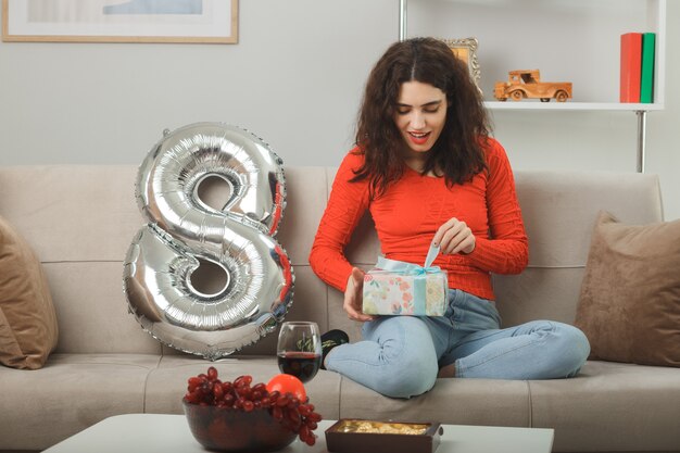 Glückliche und erfreute junge Frau in der Freizeitkleidung lächelnd fröhlich sitzend auf einer Couch mit Ballon mit der Nummer acht, die Geschenk hält, um es zu öffnen, das internationalen 8. Frauentag feiert