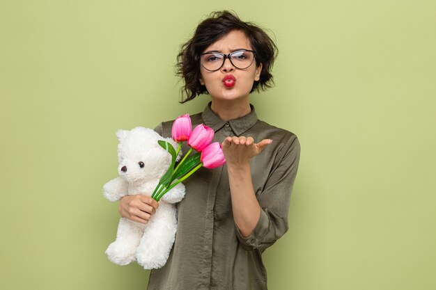 Glückliche und positive Frau mit kurzen Haaren, die einen Blumenstrauß aus Tulpen und einen Teddybären hält, der auf die Kamera schaut und einen Kuss bläst, um den internationalen Frauentag 8.