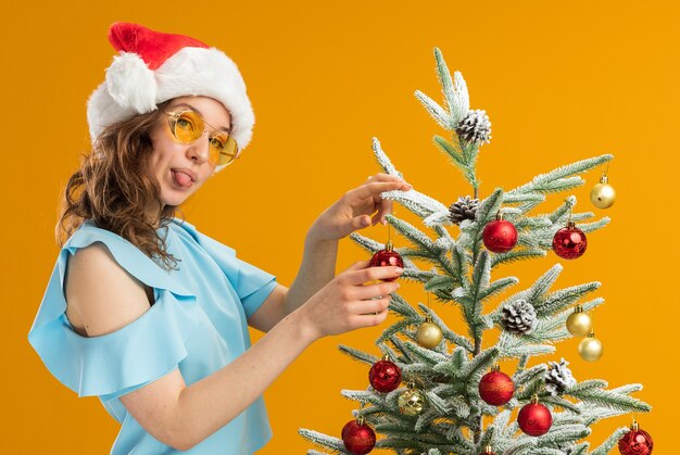 Glückliche und positive junge Frau in der blauen Spitze und in der Weihnachtsmannmütze, die gelbe Gläser tragen, die Weihnachtsbaum verzieren, der Zunge heraussteht, die über orange Hintergrund steht