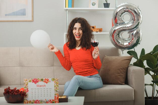 Glückliche und zufriedene junge Frau in Freizeitkleidung, die fröhlich auf einer Couch sitzt, mit einem Glas Wein, der Ballon im hellen Wohnzimmer hält, um den internationalen Frauentag am 8. März zu feiern?