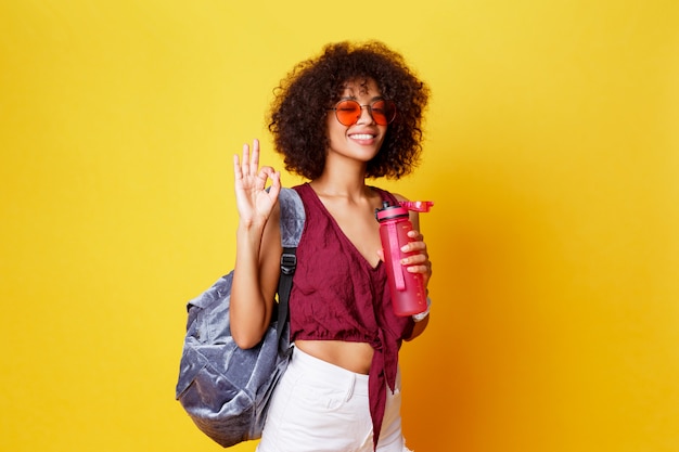 Glückliche verspielte schwarze Frau im stilvollen Sommeroutfit mit Friedenszeichen, das im Studio auf gelbem Hintergrund aufwirft. Flasche Wasser halten. Afro Frisur. Gesunder Lebensstil.