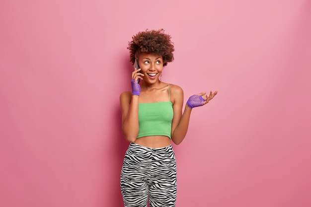 Kostenloses Foto glückliche verträumte frau mit lockigem haar genießt telefongespräch, hebt die hand und lächelt positiv, in sportkleidung gekleidet, hat schlanke figur