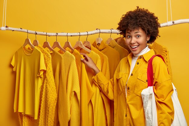 Glückliche weibliche Shopaholic wählt Kleidung auf Kleiderbügeln in eigenem Kleiderschrank, gekleidet in helle Jacke, trägt Tasche, hat ansprechendes Lächeln