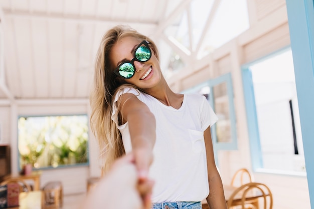 Glückliche weiße Frau mit Sonnenbräune, die in der Cafeteria aufwirft. Innenporträt des niedlichen kaukasischen weiblichen Modells mit schönen blonden Haaren.