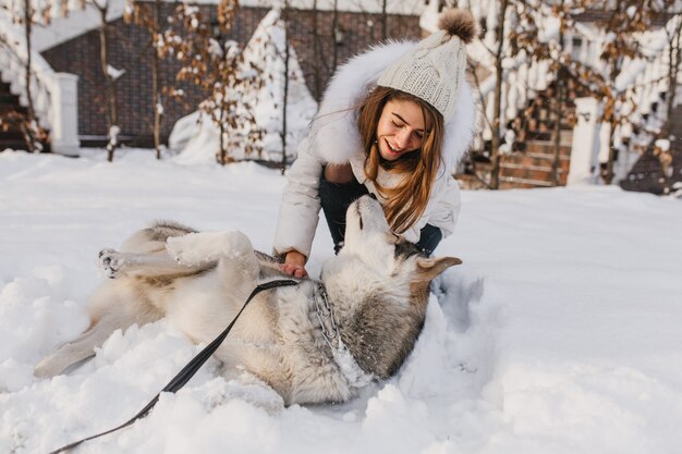 Glückliche Winterzeit der freudigen jungen Frau, die mit niedlichem husky Hund im Schnee auf Straße spielt. Fröhliche Stimmung, positive Emotionen, echte Freundschaft mit Haustieren, Liebestiere
