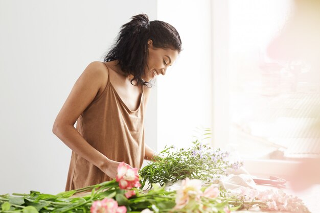 Glückliche zarte afrikanische Frau lächelnd, die Blumenstrauß am Arbeitsplatz über weißer Wand macht.