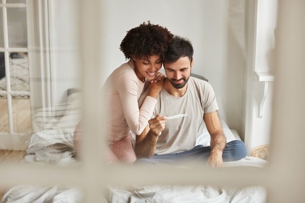 Glückliche zukünftige Eltern gemischter Rassen schauen freudig auf Schwangerschaftstest, freuen sich über positive Nachrichten über Schwangerschaft, sitzen zusammen auf dem Bett gegen häusliche Innenräume.