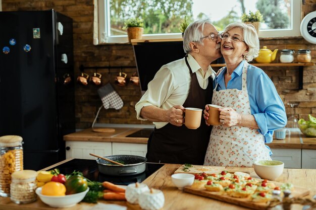 Glücklicher älterer Mann, der seine Frau küsst, während er Kaffee trinkt und mit ihr in der Küche Essen zubereitet