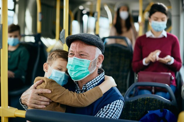 Glücklicher älterer Mann, der seinen verschlafenen Enkel hält, während er während der COVID19-Pandemie mit dem Bus pendelt
