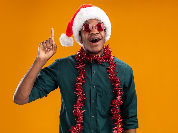Glücklicher Afroamerikanermann in der Weihnachtsmannmütze mit der Girlande, die die Brille trägt, die Zeigefinger zeigt, der neue Idee über orange Wand steht