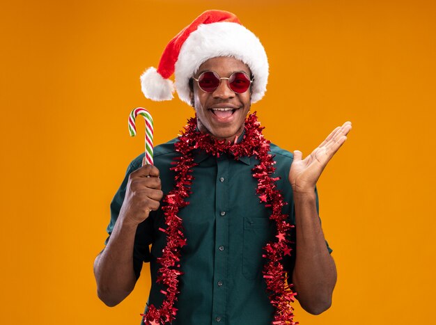 Glücklicher Afroamerikanermann in der Weihnachtsmannmütze mit der Girlande, die die Brille trägt, die Zuckerstange hält, die fröhlich mit erhöhtem Arm über orange Wand steht