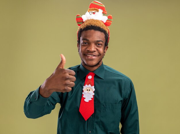 Glücklicher Afroamerikanermann mit lustigem Weihnachtsmannrand und roter Krawatte, die Kamera betrachtet, die Daumen oben lächelnd steht über grünem Hintergrund