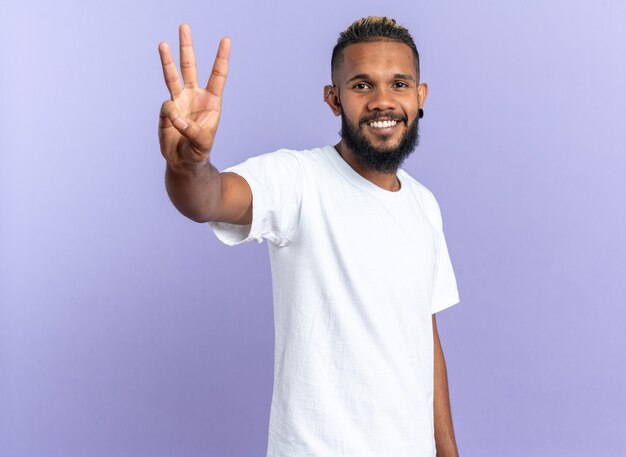 Glücklicher afroamerikanischer junger Mann im weißen T-Shirt, der die Kamera anschaut und mit den Fingern Nummer drei lächelnd nach oben zeigt