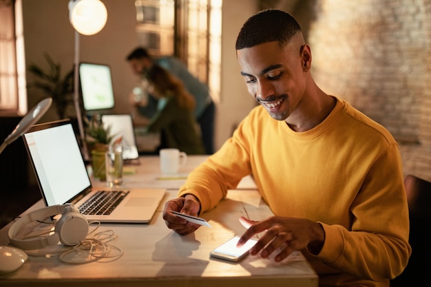 Glücklicher afroamerikanischer Unternehmer, der Handy und Kreditkarte von Online-Zahlungen verwendet, während er nachts im Büro arbeitet