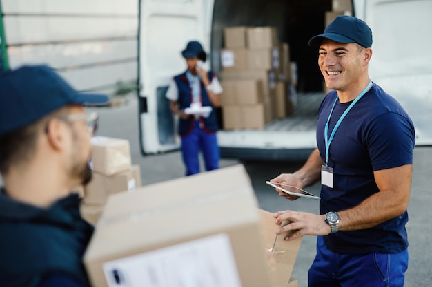 Glücklicher Arbeiter, der Touchpad verwendet, während er mit seinem Kollegen kommuniziert und die Paketzustellung organisiert