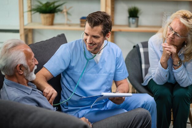 Glücklicher Arzt, der den Herzschlag eines reifen Patienten hört, während er seinen während eines Hausbesuchs untersucht