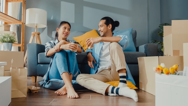 Glücklicher asiatischer junger Paarmann und -frau sitzen am neuen Haus, trinken Kaffee und sprechen mit Kartonverpackungsbox-Lagerung