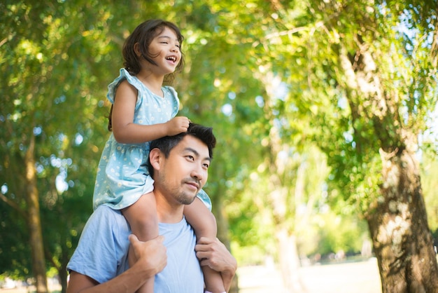 Glücklicher asiatischer Vater mit Tochter am Hals. Glücklicher Mann, der Ruhe im Park genießt, und kleines Mädchen, das auf seinen Schultern sitzt und beiseite schaut. Familiensommeraktivität, Ruhe und glückliche Momente Konzept