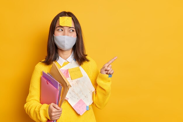 Glücklicher asiatischer weiblicher Manager, der beschäftigt ist, Papierarbeit zu tun, trägt schützende Einwegmaske während der Coronavirus-Pandemie zeigt Zeigefinger auf Kopierraum an