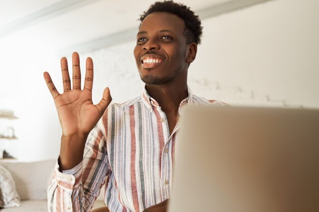 Glücklicher attraktiver junger afroamerikanischer männlicher Blogger, der Bilder hochlädt, neuen Beitrag für soziale Netzwerke tippt, mit seinen Anhängern online plaudert, im Café sitzt, Hand winkt und breit lächelt