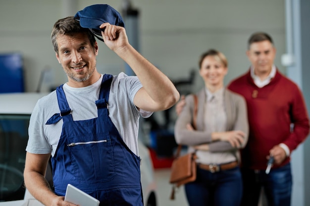 Glücklicher Automechaniker, der seinen Hut nimmt, während er in einer Werkstatt steht und in die Kamera schaut Seine Kunden sind im Hintergrund