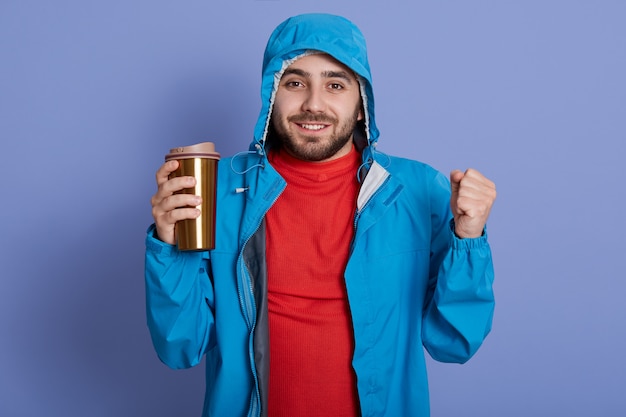 Glücklicher bärtiger Mann, der blaue Jacke und rotes Hemd trägt und heißes Getränk vom Thermobecher und geballten Fäusten genießt,