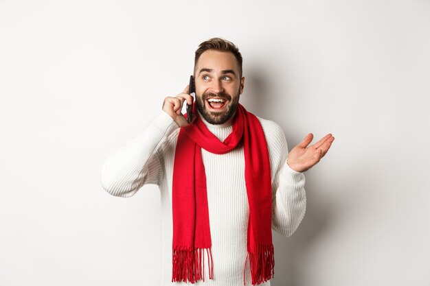 Glücklicher bärtiger Mann, der frohe Weihnachten am Telefon wünscht, jemanden anruft und spricht, im Pullover mit rotem Schal, weißer Hintergrund steht.