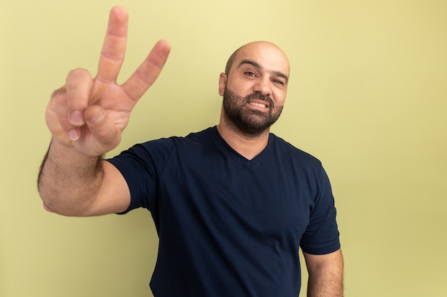 Kostenloses Foto glücklicher bärtiger mann im schwarzen t-shirt, der zuversichtlich lächelt und v-zeichen zeigt, das über grüner wand steht