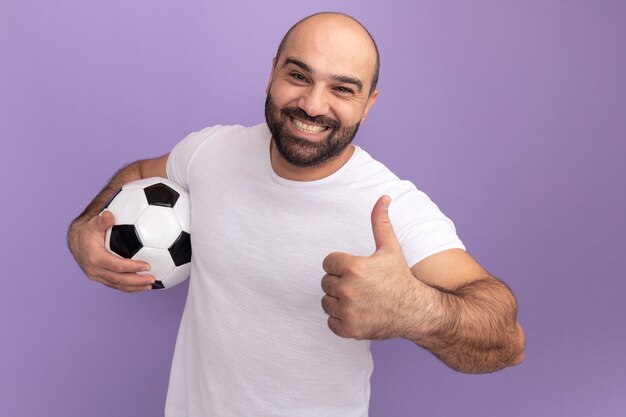 Glücklicher bärtiger Mann im weißen T-Shirt, der Fußball lächelnd fröhlich zeigt Daumen hoch stehend über lila Wand