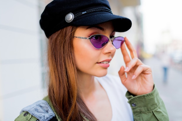 Glücklicher brünetter Weoman, der im Freien aufwirft. Stilvoller Hut, Sonnenbrille und grüne Jacke.