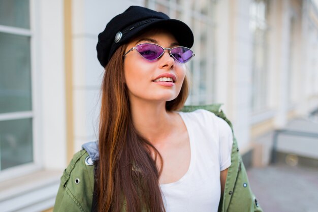 Glücklicher brünetter Weoman, der im Freien aufwirft. Stilvoller Hut, Sonnenbrille und grüne Jacke.