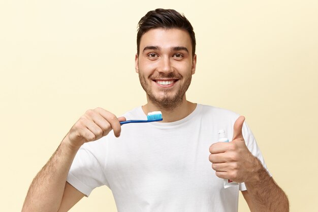 Glücklicher energischer junger europäischer Kerl mit Stoppeln, die Zahnbürste mit Bleichpaste halten und Daumen hoch Geste zeigen, die in guter Stimmung ist.