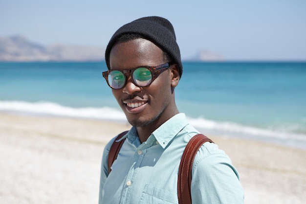 Kostenloses Foto glücklicher entspannter junger dunkelhäutiger hipster in den schatten und in der kopfbedeckung, die gegen klaren blauen himmel und weites azurblaues meer stehen, sommerferien im ferienort verbringen und mit freudigem lächeln schauen