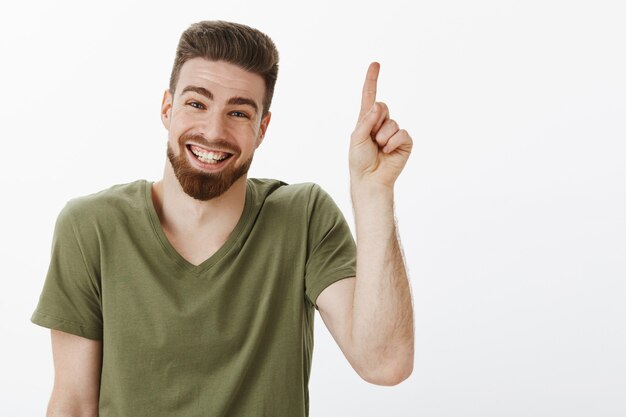 Glücklicher entzückter und sorgloser attraktiver erwachsener Mann mit Bart, der freudig lachend große Zeit hat, entzückt und freudig aussehend zeigt oder Nummer eins über weißer Wand zeigt