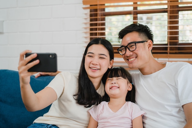 Glücklicher fröhlicher asiatischer Familienvater, Mutter und Kinder, die Spaß haben und Smartphone-Videoanruf auf Sofa im Haus verwenden.