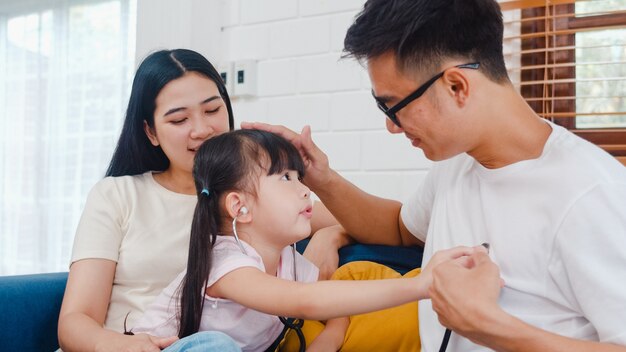 Glücklicher fröhlicher asiatischer Familienvater, Mutter und Tochter, die lustiges Spiel als Arzt spielen, der Spaß auf Sofa zu Hause hat
