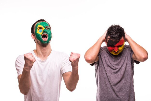 Glücklicher Fußballfan von Brasilien feiern Sieg über verärgerten Fußballfan der deutschen Nationalmannschaften mit gemaltem Gesicht lokalisiert auf weißem Hintergrund