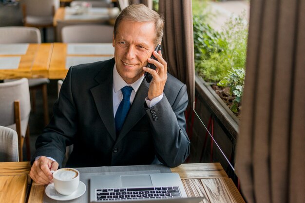 Glücklicher Geschäftsmann, der am Handy mit Tasse Kaffee und Laptop auf Tabelle spricht