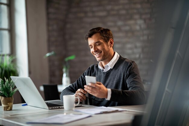 Glücklicher Geschäftsmann, der auf dem Handy SMS schreibt, während er im Büro am Laptop arbeitet