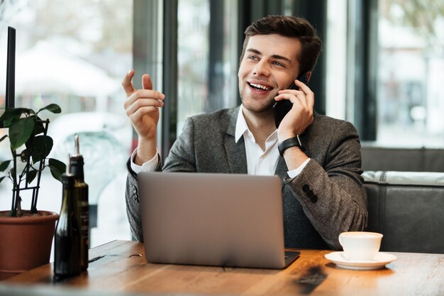 Glücklicher Geschäftsmann, der durch die Tabelle im Café mit Laptop-Computer bei der Unterhaltung durch Smartphone sitzt