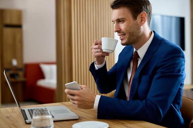 Glücklicher Geschäftsmann, der eine Tasse Kaffee trinkt, während er E-Mails auf dem Laptop im Hotelzimmer liest