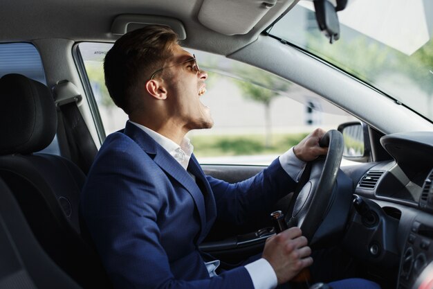 Glücklicher Geschäftsmann sitzt Shows seine Gefühle, die am Lenkrad innerhalb des Autos sitzen