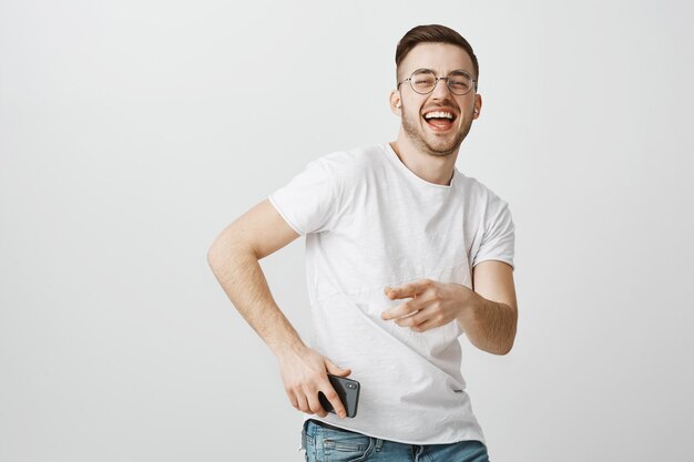 Glücklicher gutaussehender Kerl in den Gläsern, die zur Musik in drahtlosen Kopfhörern mit Handy in der Hand tanzen