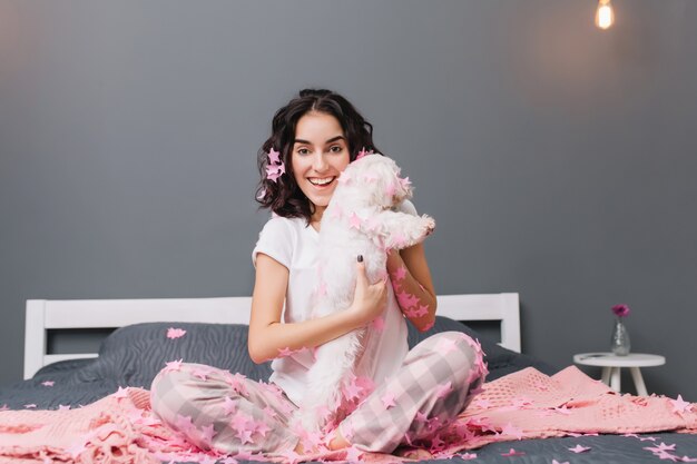 Glücklicher guter Morgen, wahre positive Gefühle der jungen freudigen Frau im Pyjama mit brünettem lockigem Haar, das Spaß mit kleinem Hund in rosa Lametta auf Bett hat