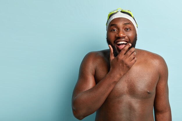 Glücklicher hemdloser Mann mit dunkler Haut, genießt Schwimmen, Erholungszeit und Fitness, hält Kinn, schaut positiv