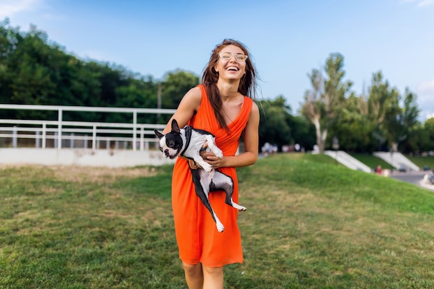 Glücklicher hübscher Frauenpark, der Boston-Terrier-Hund hält, positive Stimmung lächelnd, trendiger Sommerstil, orange Kleid, Sonnenbrille tragend, mit Haustier spielend, Spaß, bunt