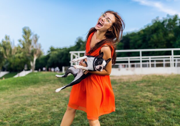Glücklicher hübscher Frauenpark, der Boston-Terrier-Hund hält, positive Stimmung lächelnd, trendiger Sommerstil, orange Kleid, Sonnenbrille tragend, mit Haustier spielend, Spaß, bunt